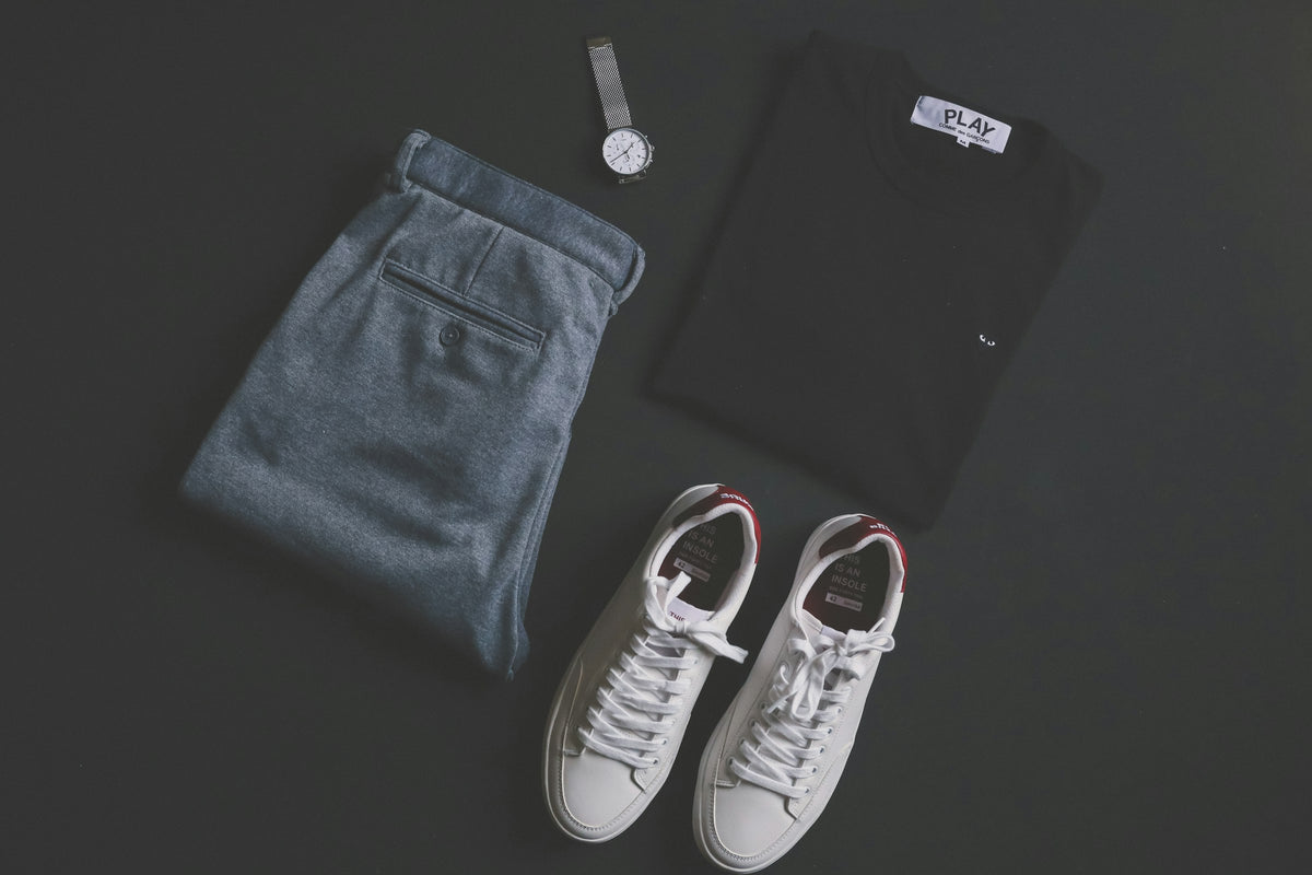 Flat lay of white low-top sneakers, folded trousers, a black T-shirt and a watch