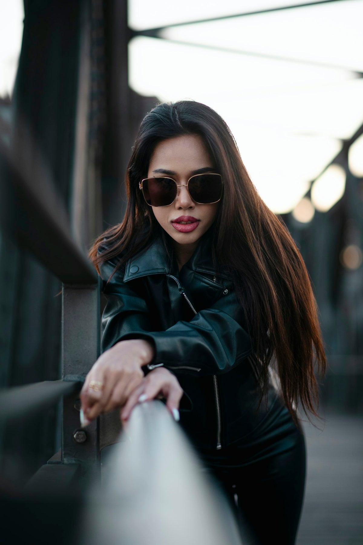 Woman in sunglasses and a black leather jacket leaning on a bridge railing